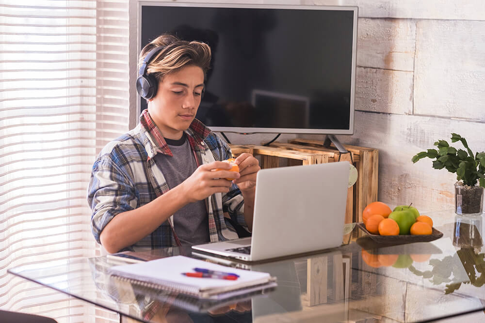 teen eats a healthy snack after school while doing homework on his laptop